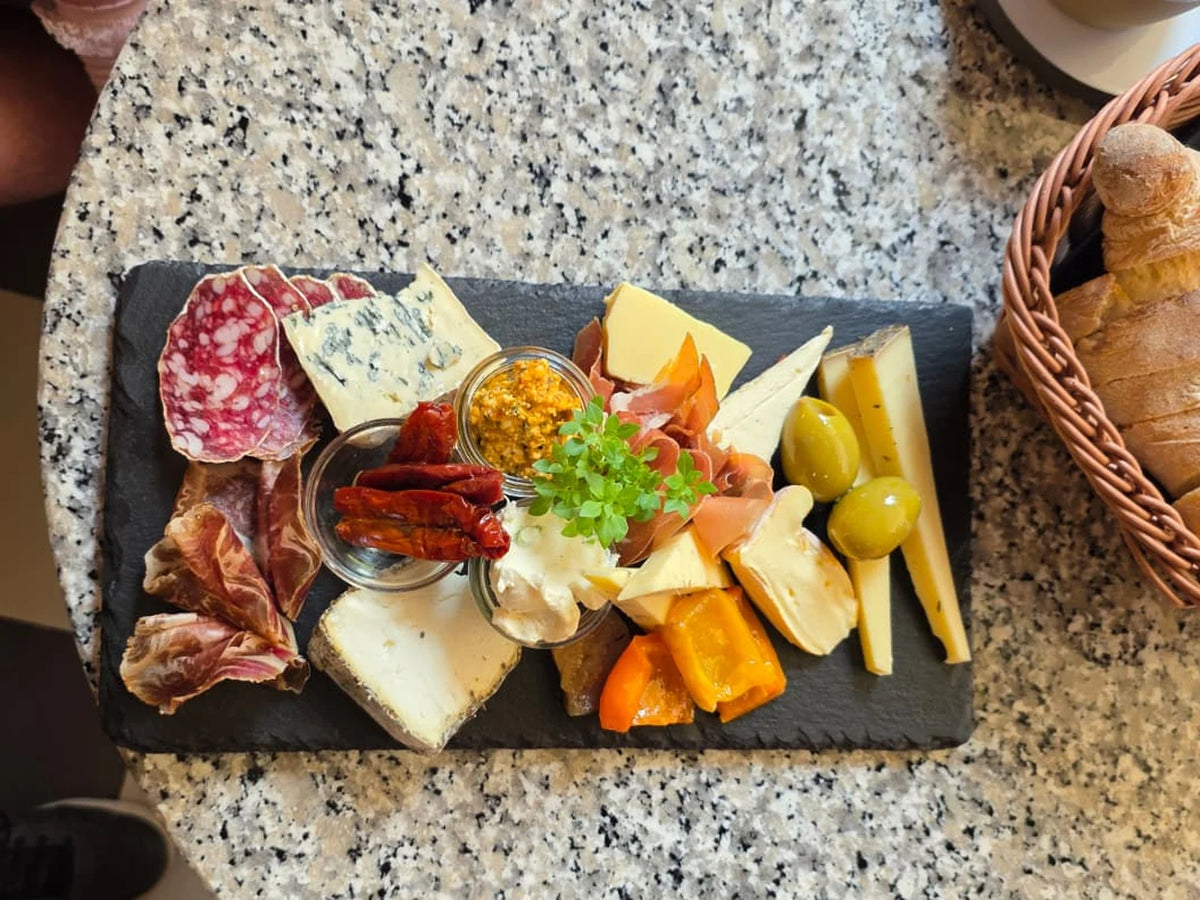 Käseplatte mit Schinken, Oliven, Feigensenf und Antipasti auf Schiefer, daneben der Brotkorb