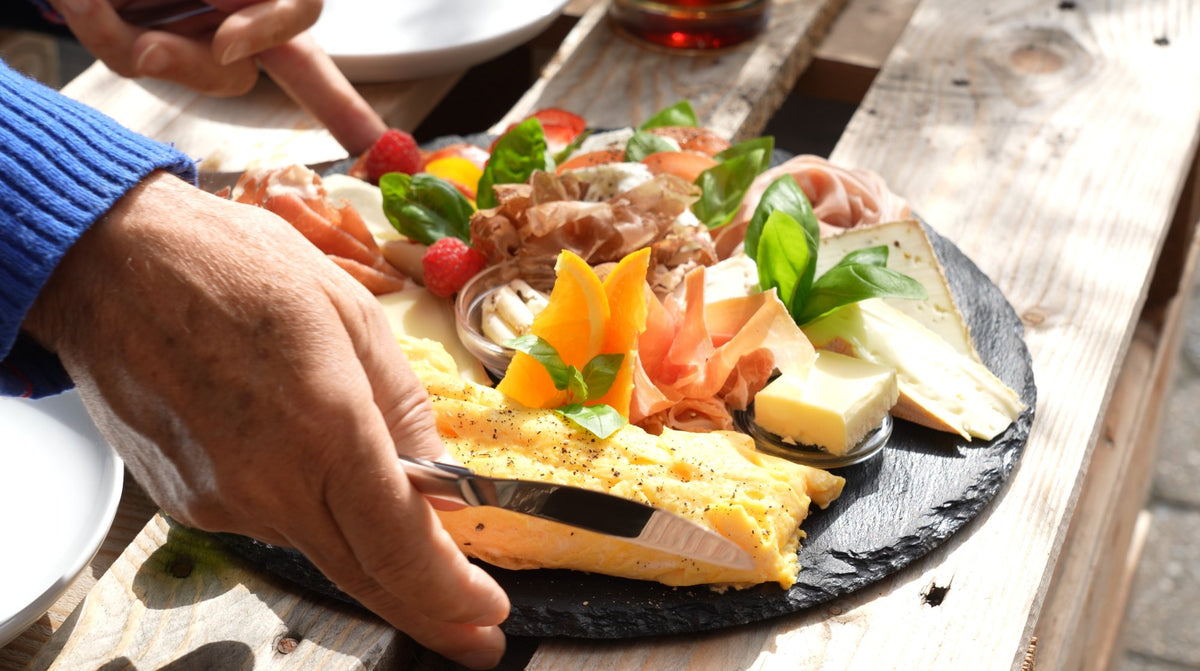 Gast greift zu einer angerichteten Käse- und Schinkenplatte auf Schiefer an einem Holztisch