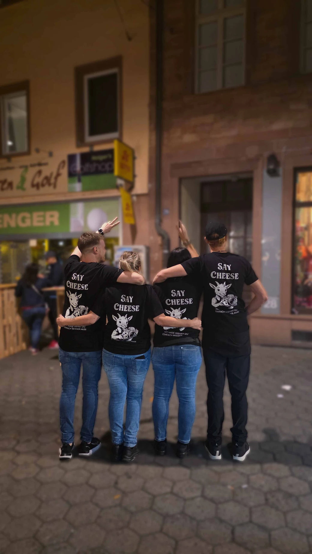 Vier Leute aus dem Team stehen abends Arm in Arm in der Sandgasse, auf den Shirts steht Say Cheese.
