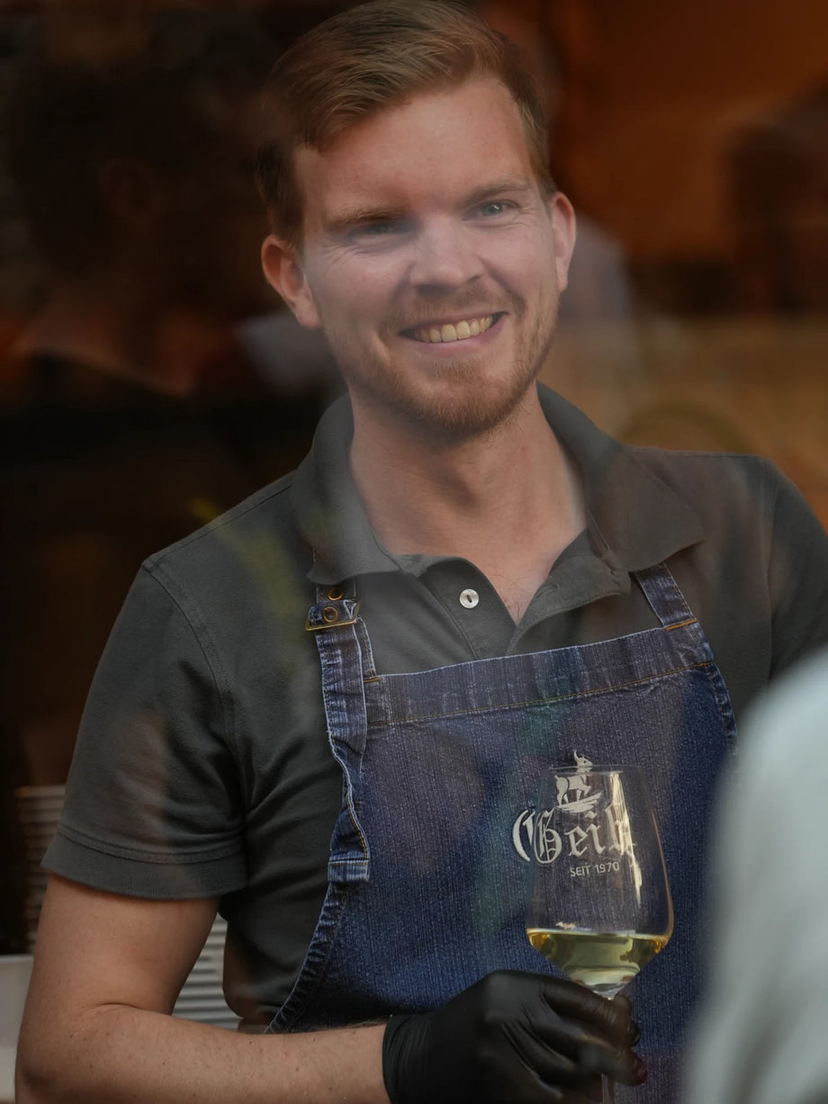 Manu lächelt hinter der Theke, ein Glas Weißwein in der Hand.