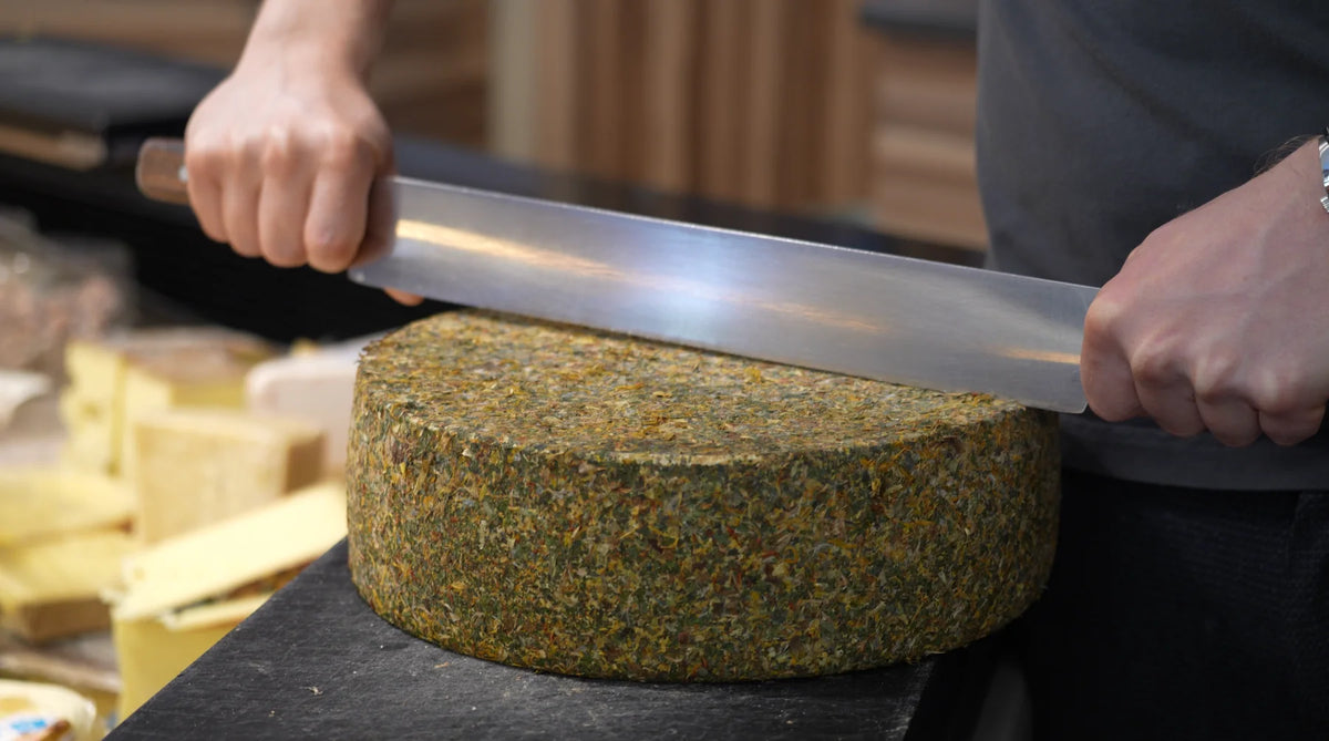 Ein Kräuterkäse wird von Hand mit dem großen Käsemesser aufgeschnitten