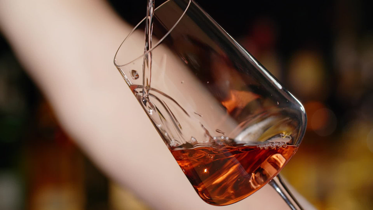 Wein wird in ein geneigtes Glas eingeschenkt, im Hintergrund die unscharfe Flaschenwand einer Bar